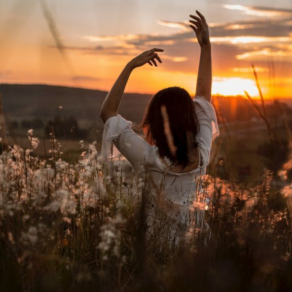 woman-wearing-white-dress-raising-her-two-hands-surrounded-white-petaled-flowers-during-sunset